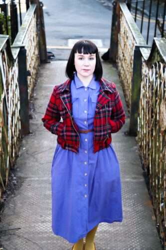 Debbie armour standing in a close in Glasgow wearing a tartan jacket and a blue dress with black bob haircut